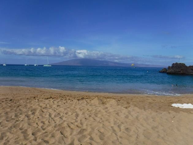 Black Rock Beach - Kaanapali. Sand in half of the photo with the ocean and another island on the other half. Sailboats are floating in the water. 