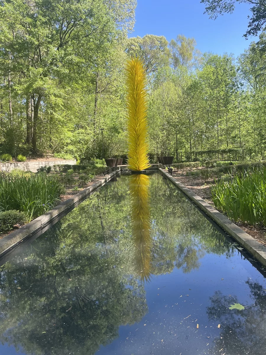 Yellow sculpture at the end of a long pond. Trees are surrounding both. 