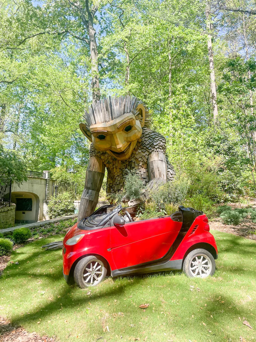 Giant wooden troll with red car, surrounded by greenery. 
