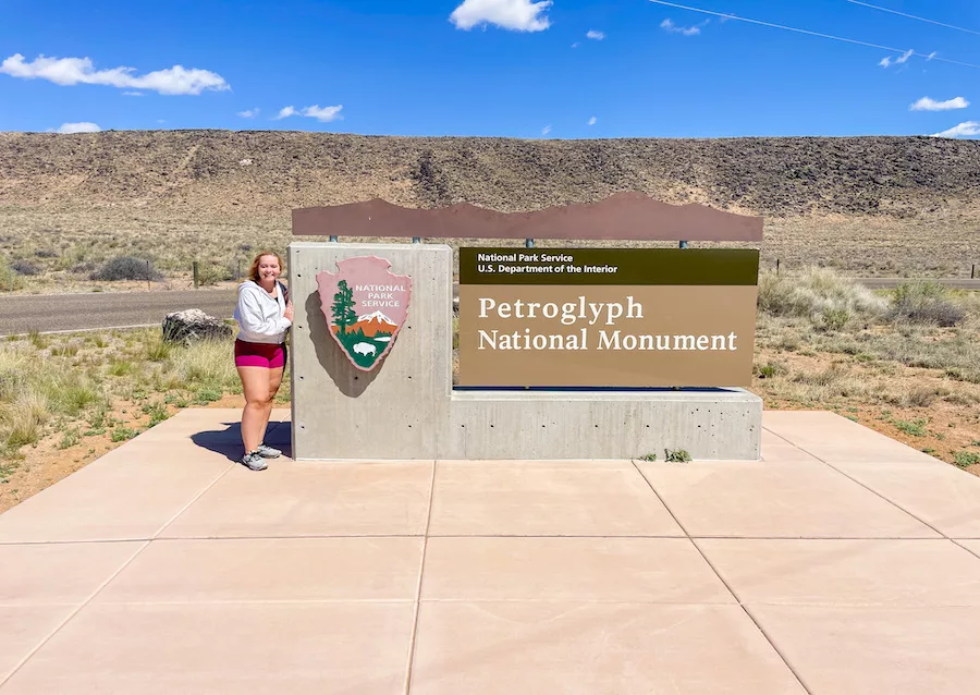 One Day in Albuquerque - Young Woman standing next to entrance sign for Petroglyph National Monument with mountains in background.