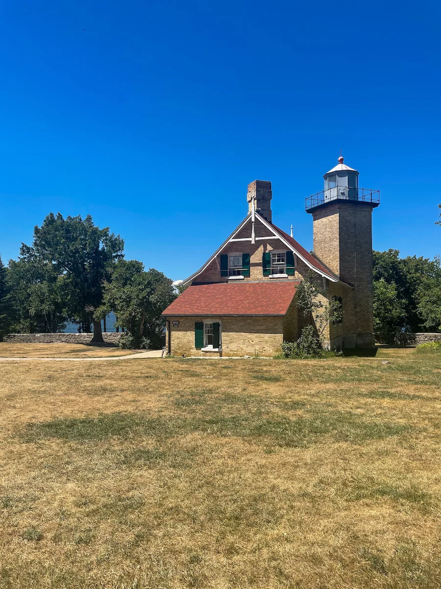 Eagle Bluff Lighthouse surrounded by grass and trees, with a bright blue sky and lake Michigan in the background.