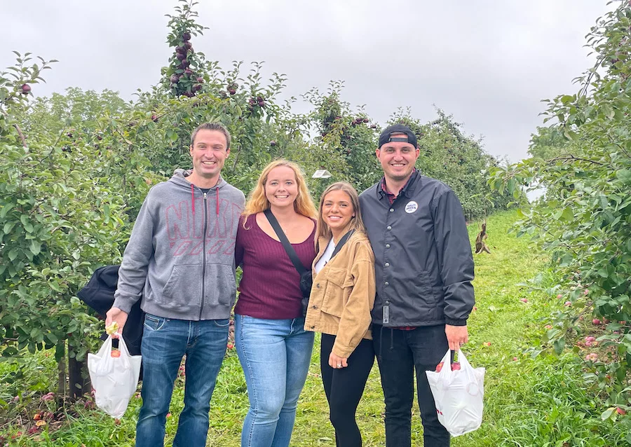 Two couples posing for a photo with two bags of apples in an apple orchard after apple picking