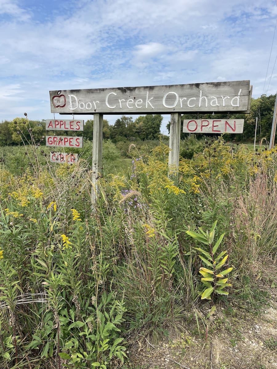 Image of Welcome Sign to Door Creek Orchards, surrounded by tall prairie grasses and wildflowers. Blue sky with clouds. 