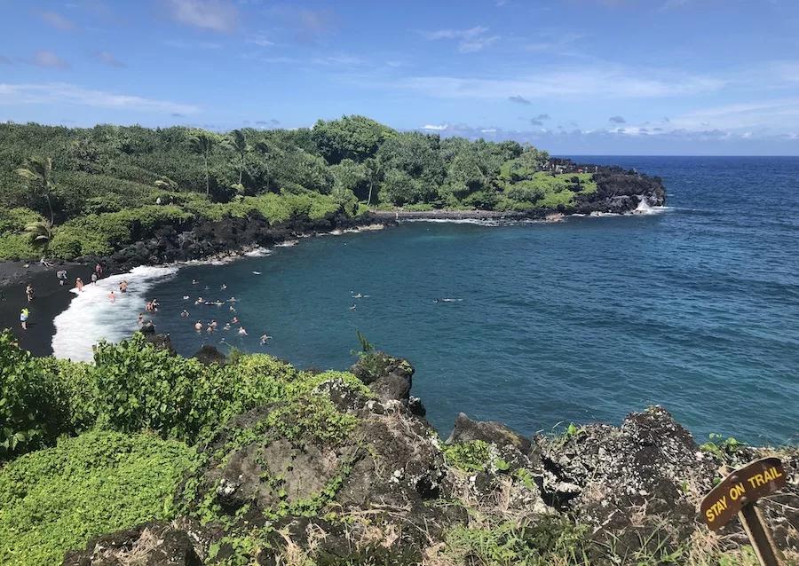 Maui Itinerary Post - Deep blue ocean water surrounded by a black sand beach and green foliage.