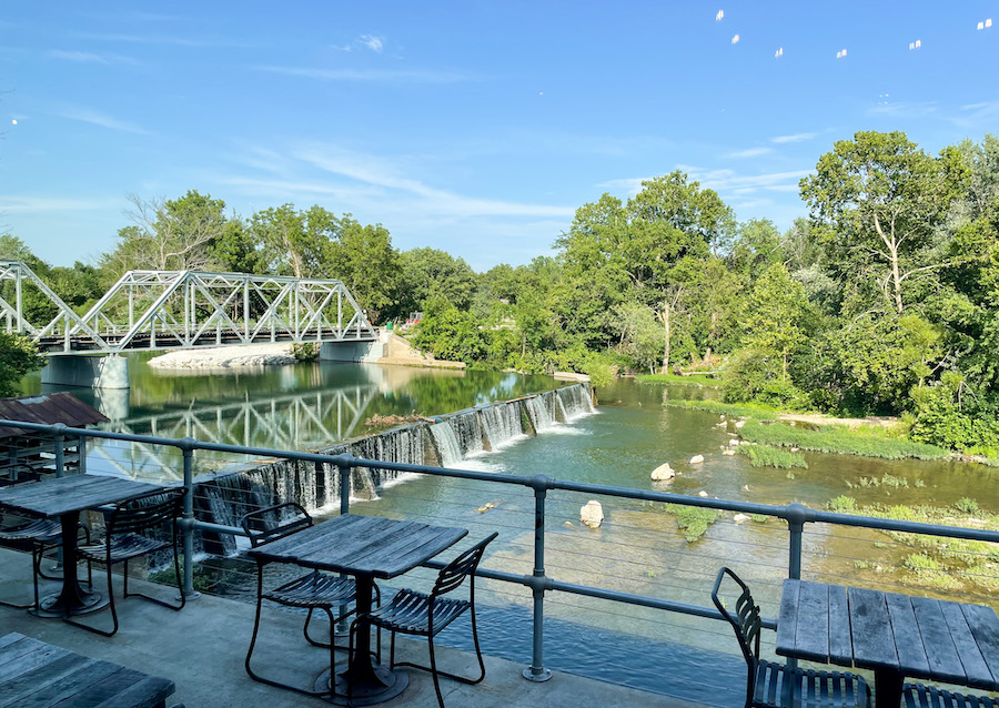 Image of beautiful river, small waterfall and white bridge from restaurant patio. Surrounded by blue sky and full green trees. 