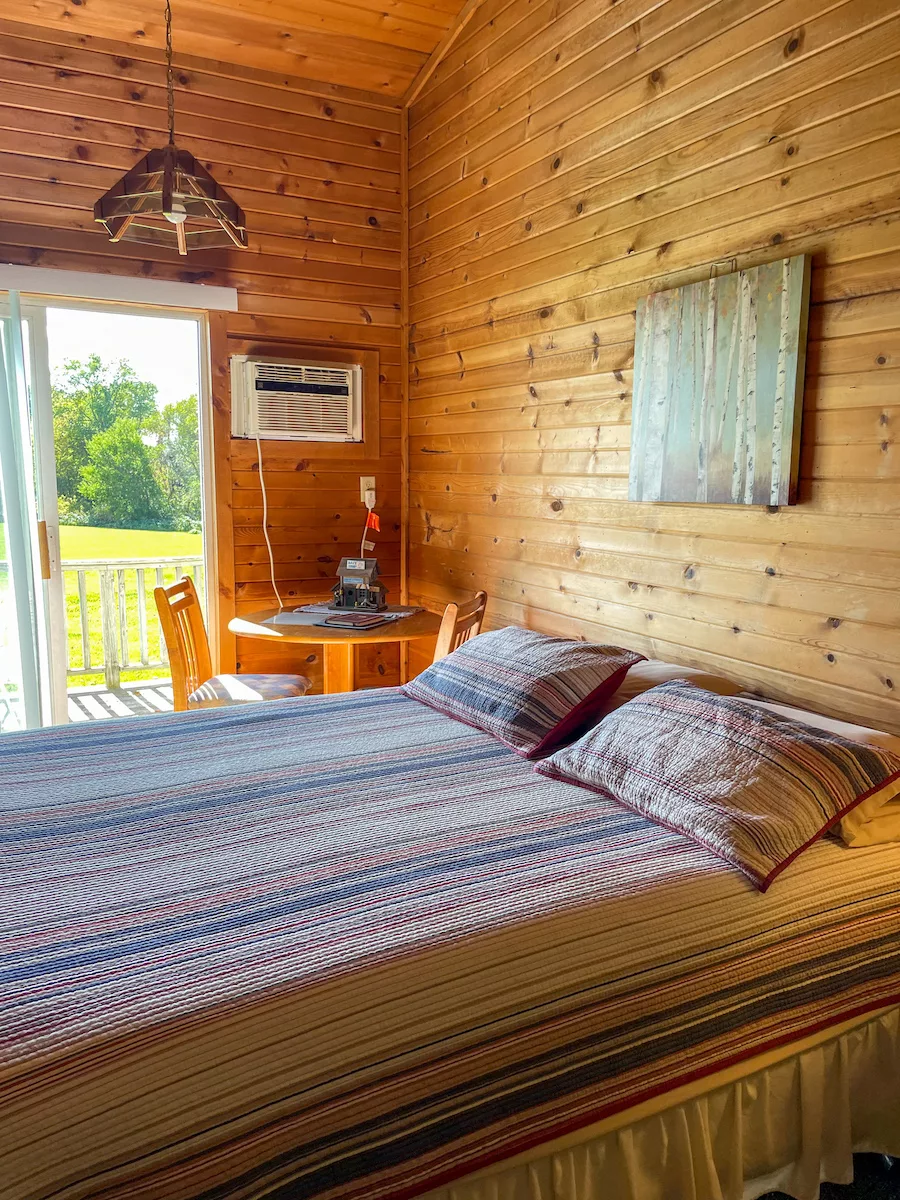 Interior room of wood cabin with bed and small kitchen table and a sliding glass door opening to a deck - at Hickory Hideaway Cabins