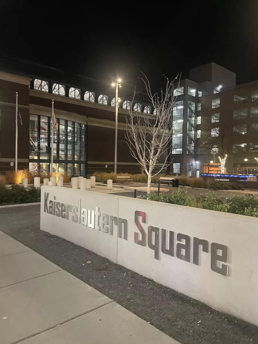 Entrance sign to Kaiserslautern Square at night with buildings lit up behind it - Downtown Davenport 