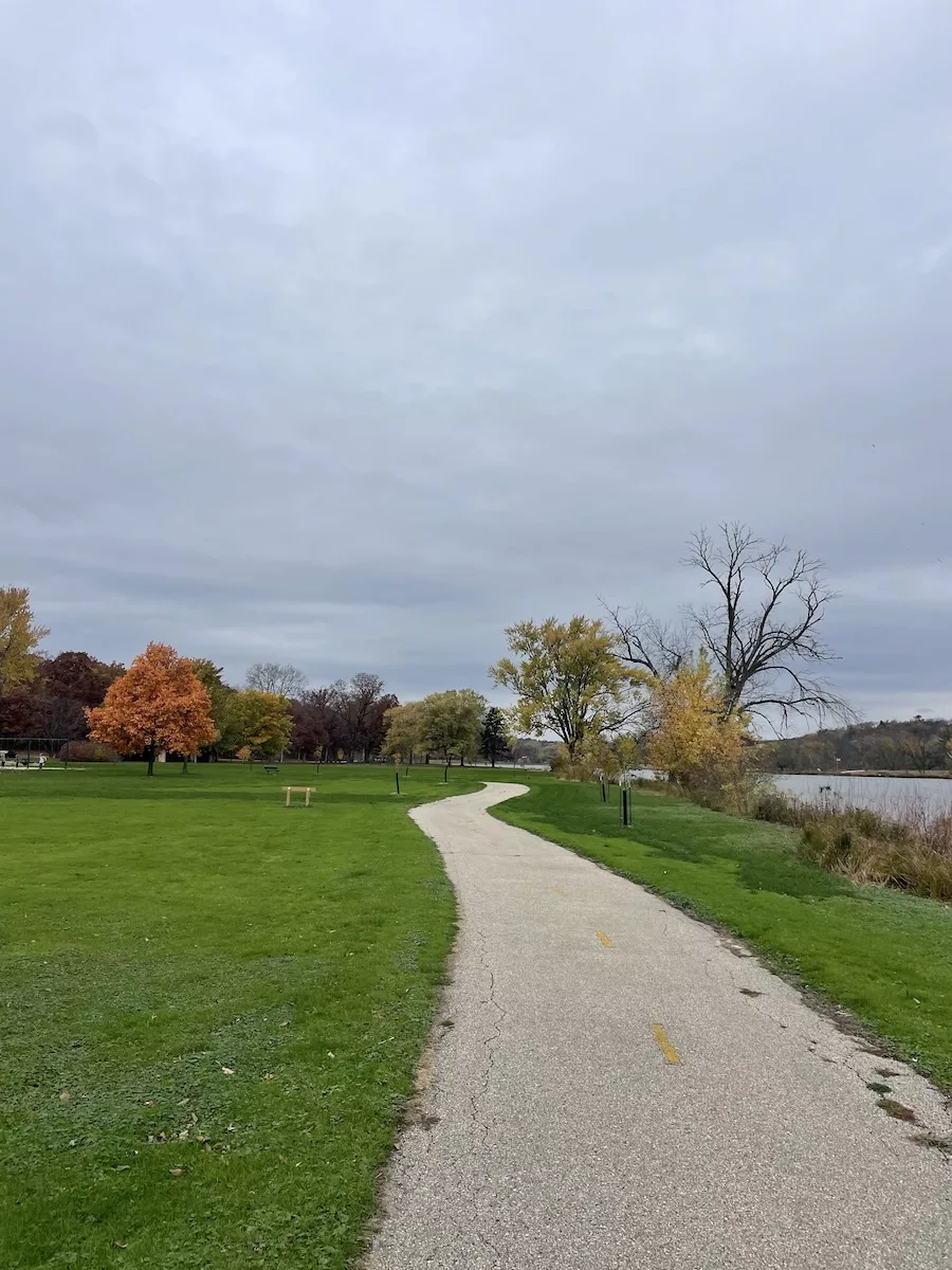 Image of paved path surrounded by green grass and fall-colored trees - Janesville Segment Ice Age Trail