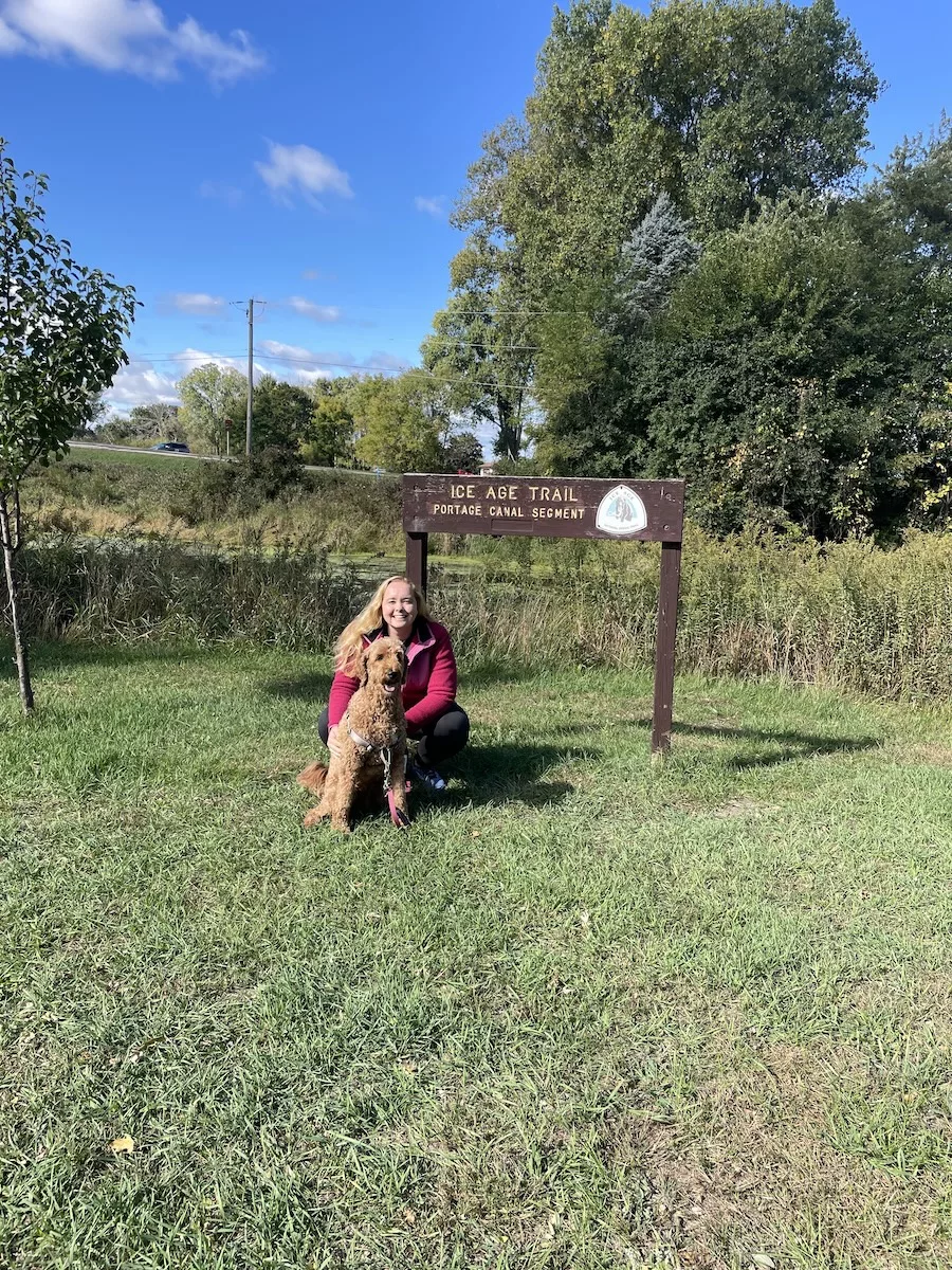 Woman and dog in front of Ice Age Trail segment sign with trees and blue sky in background