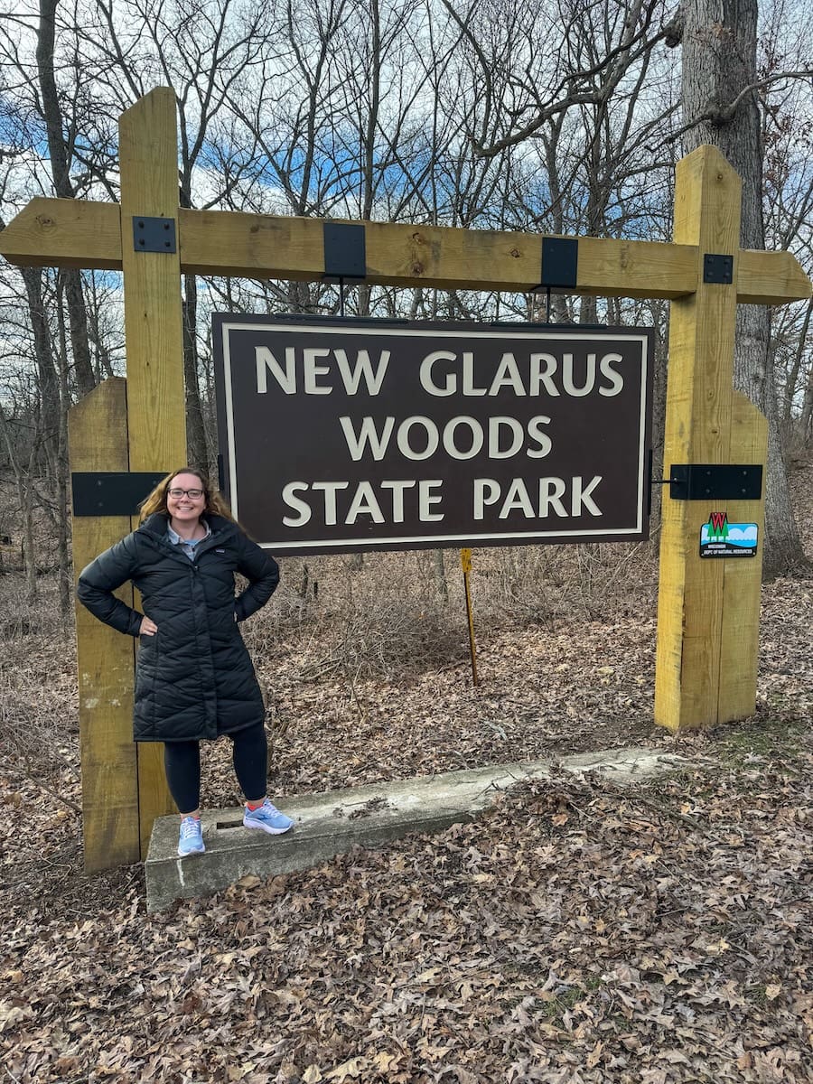 Image of a young woman in front of a large wooden sign to New Glarus Woods State Park - Guide to New Glarus