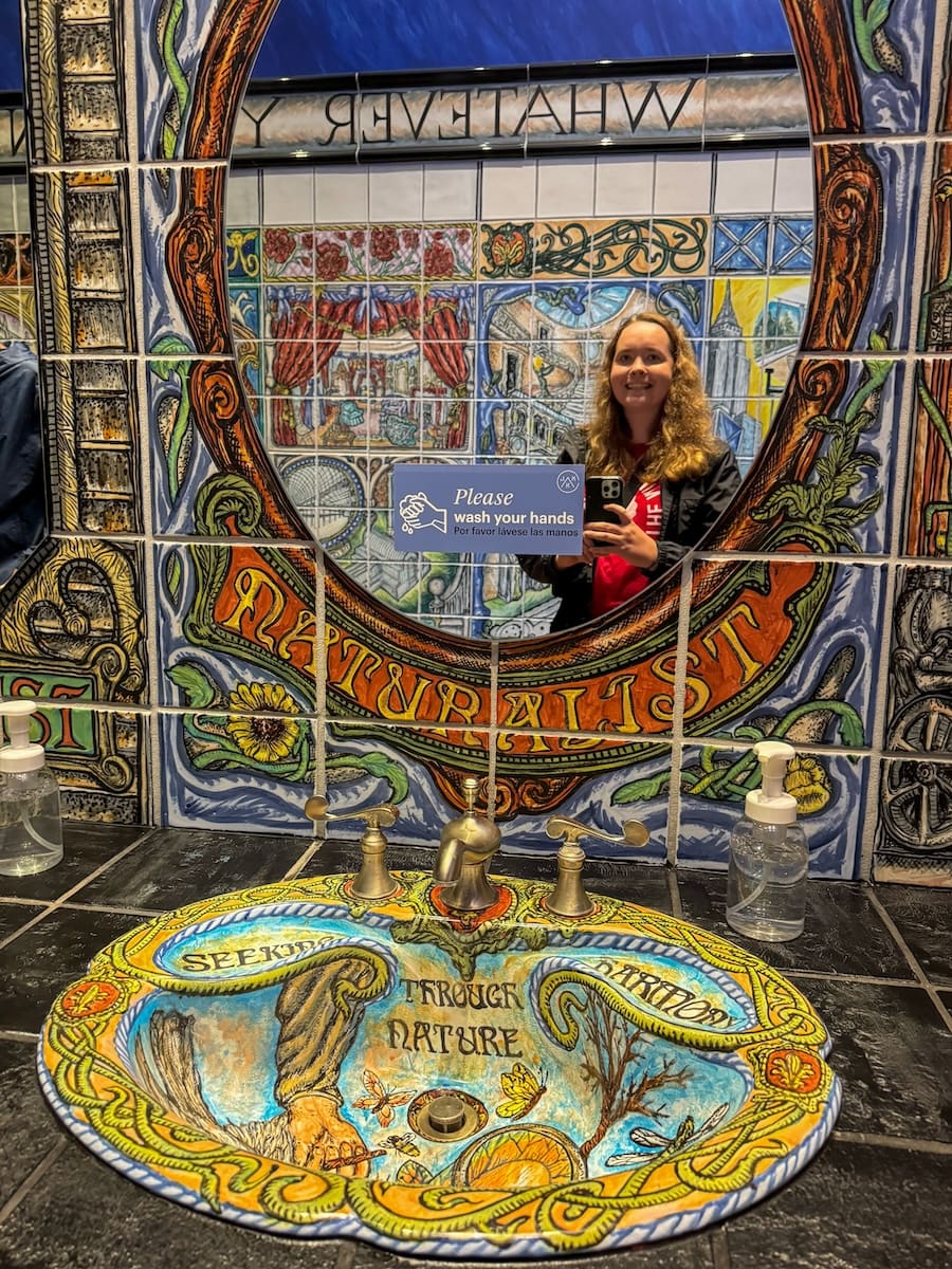 Image of colorful and artistic bathroom sin and shower area, with young woman smiling in the mirror as she takes a picture. 