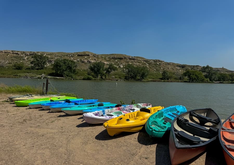 Image of blue sky and slightly green water with a row of multi-colored kayaks lining the sand and water. 