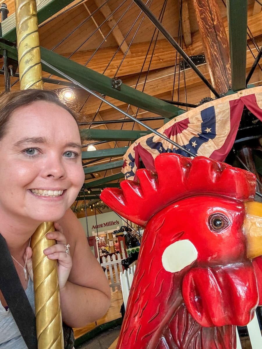 A selfie photograph of a young lady riding a rooster animal on a carousel in Shipshewana, Indiana