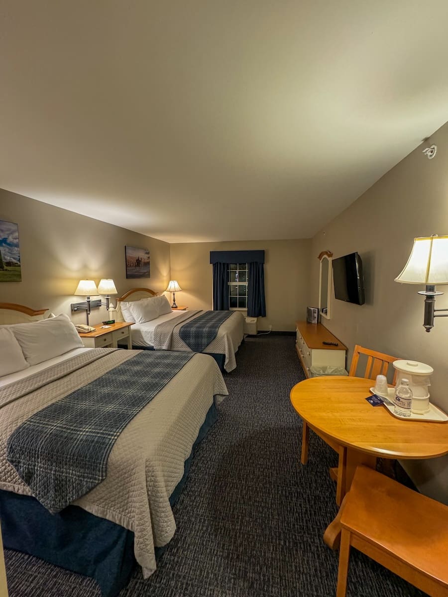 Image of a hotel room with two beds, several lamps, a tv, and a small dining room table. 