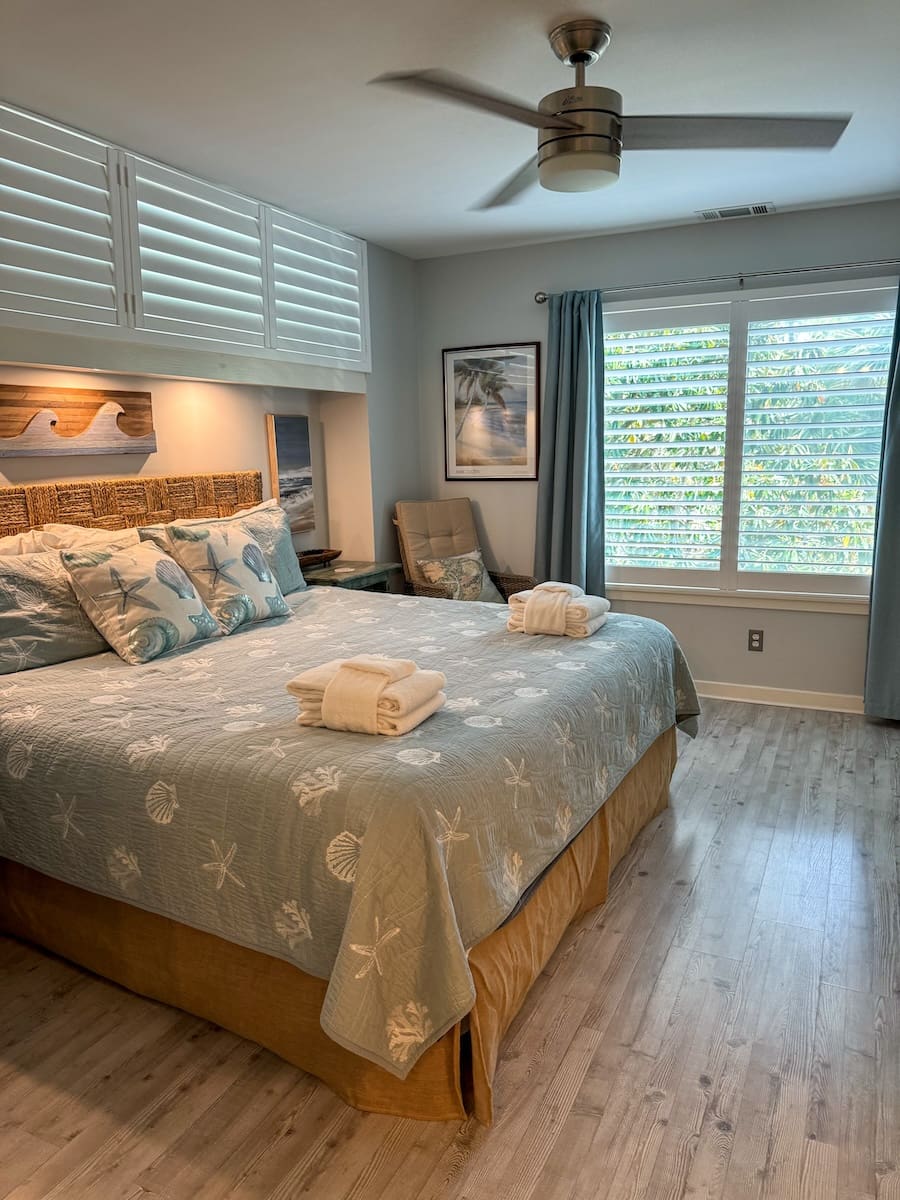 Image of a master bedroom with a king-sized bed made with a quilted comforter, large shaded window, and ceiling fan. 