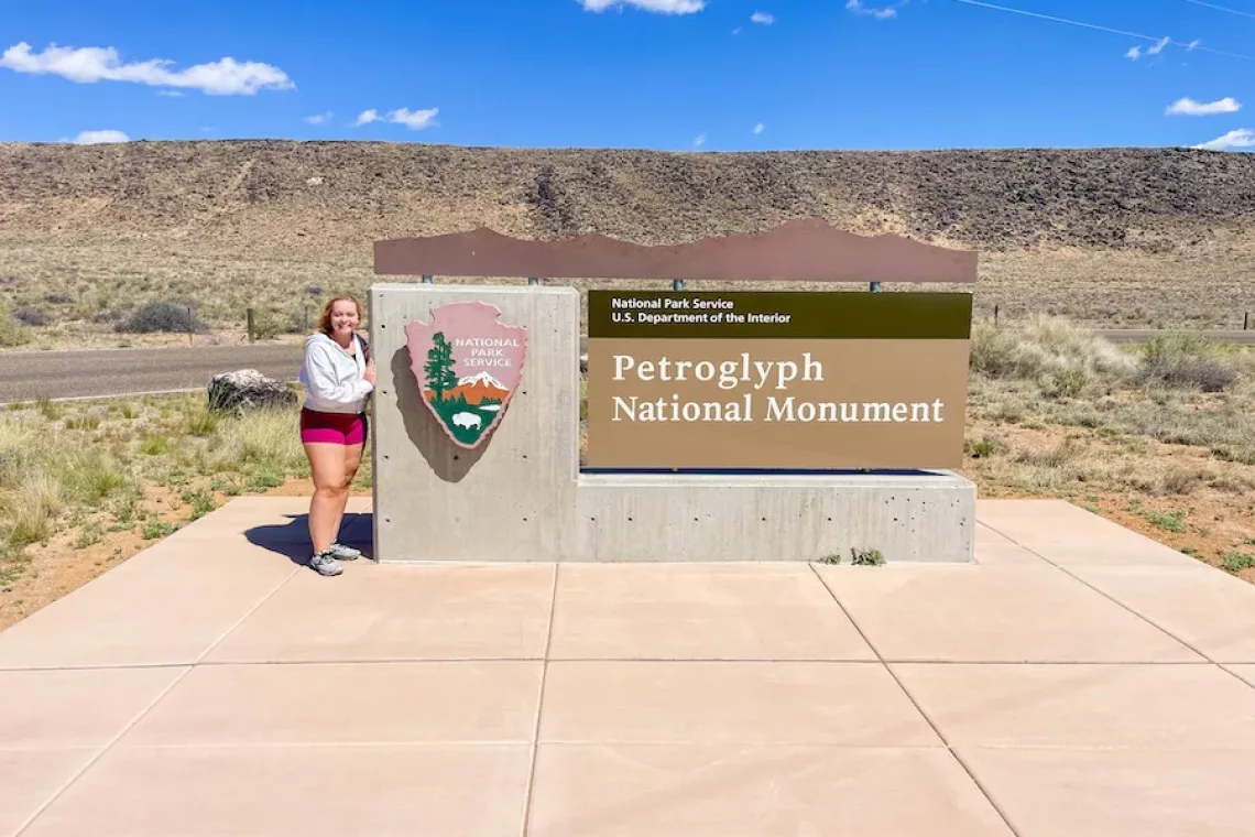 One Day in Albuquerque - Young Woman standing next to entrance sign for Petroglyph National Monument with mountains in background.