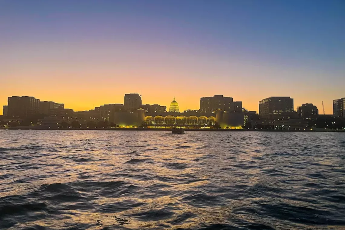 View of downtown Madison, WI at sunset with the capitol and other buildings in the background, surround by a lake.