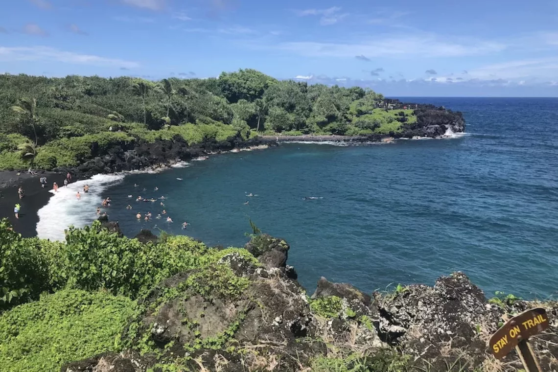 Maui Itinerary Post - Deep blue ocean water surrounded by a black sand beach and green foliage.